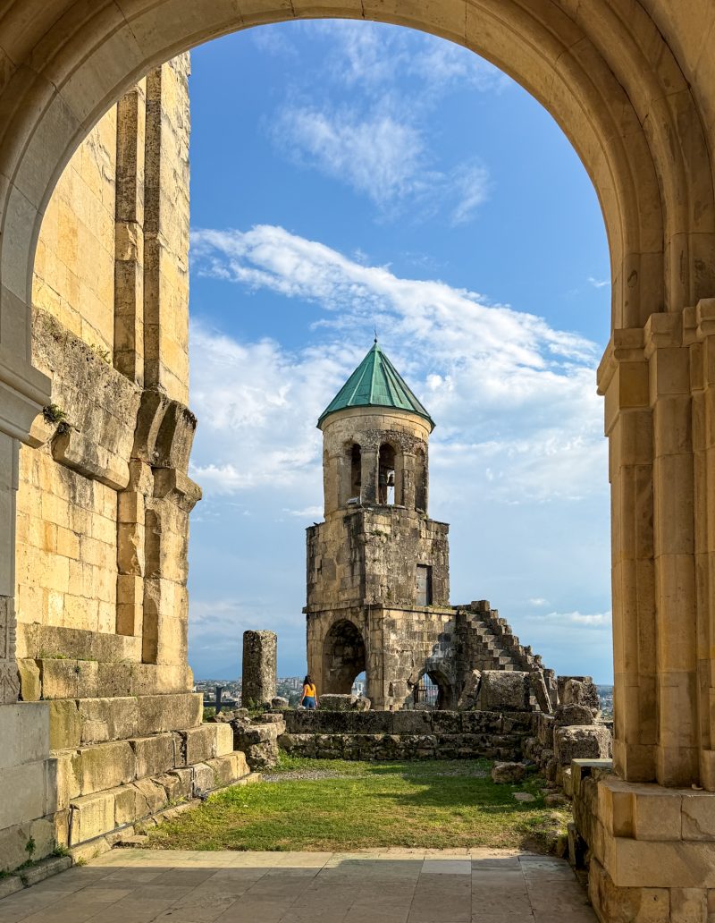 Ruined bell tower of Bagrati Cathedral seen through a stone arch, with Kutaisi in the background